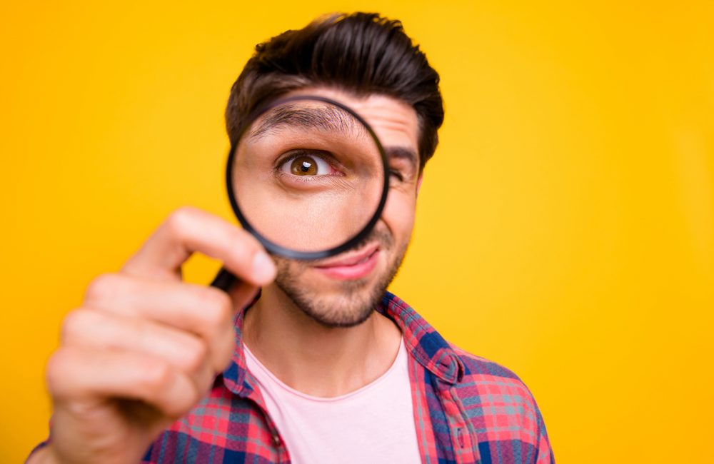 Photo of amazed man trying, to see microorganisms with loupe but he evidently cannot manage to do it while isolated with yellow background